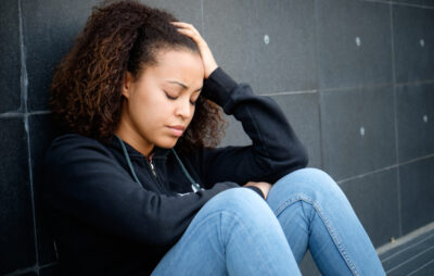 A depressed teenager sitting on the floor with her back leaning against the wall and her hand to her head